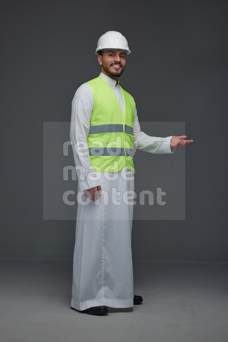 A Saudi man wearing Thobe with a yellow safety vest and white helmet standing and pointing different angles eye level on a gray background