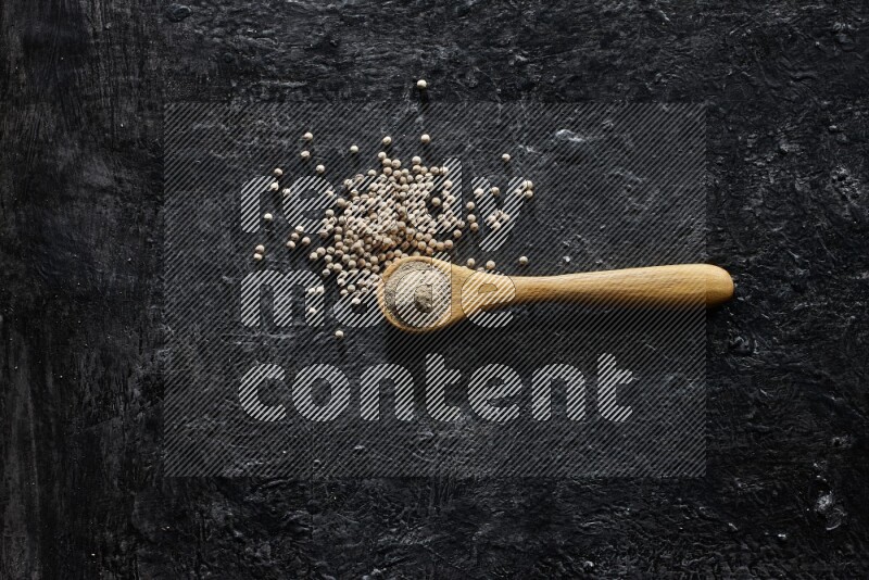 A wooden spoon full of white pepper powder with white pepper beads on textured black flooring