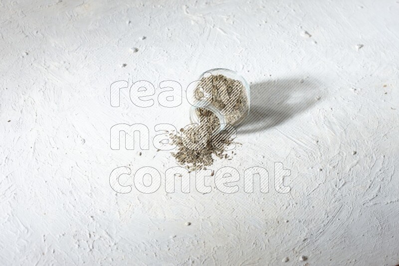 A flipped glass spice jar full of cumin seeds and the seeds spilled out on textured white flooring