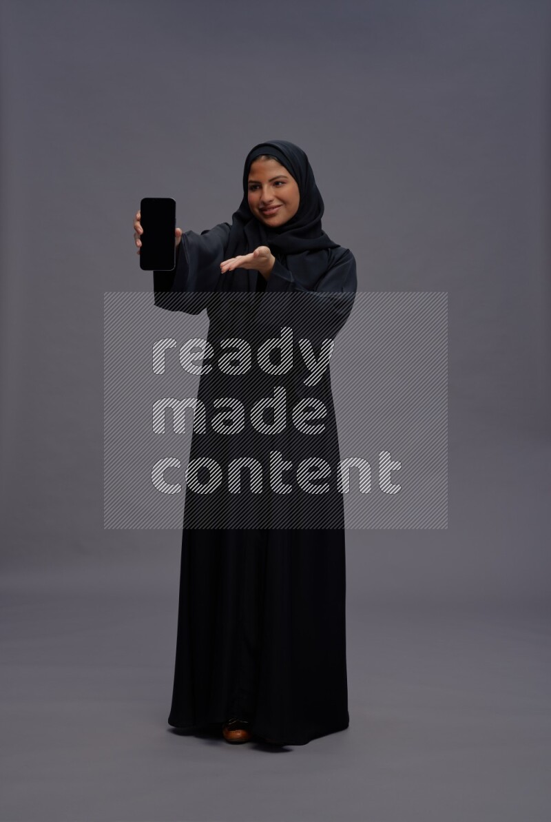 Saudi woman wearing Abaya standing showing phone to camera on gray background