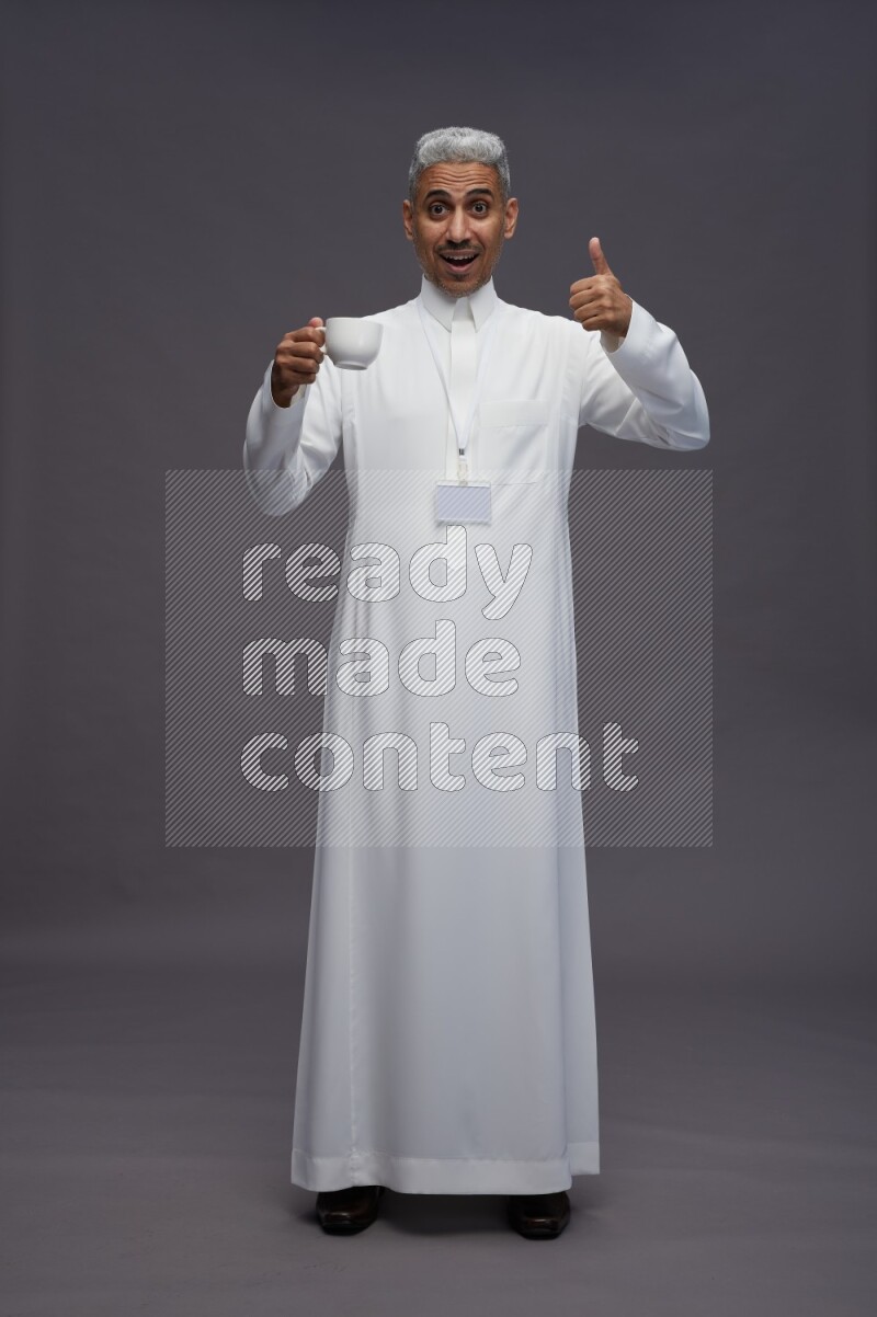 Saudi man wearing thob with neck strap employee badge standing holding mug on gray background
