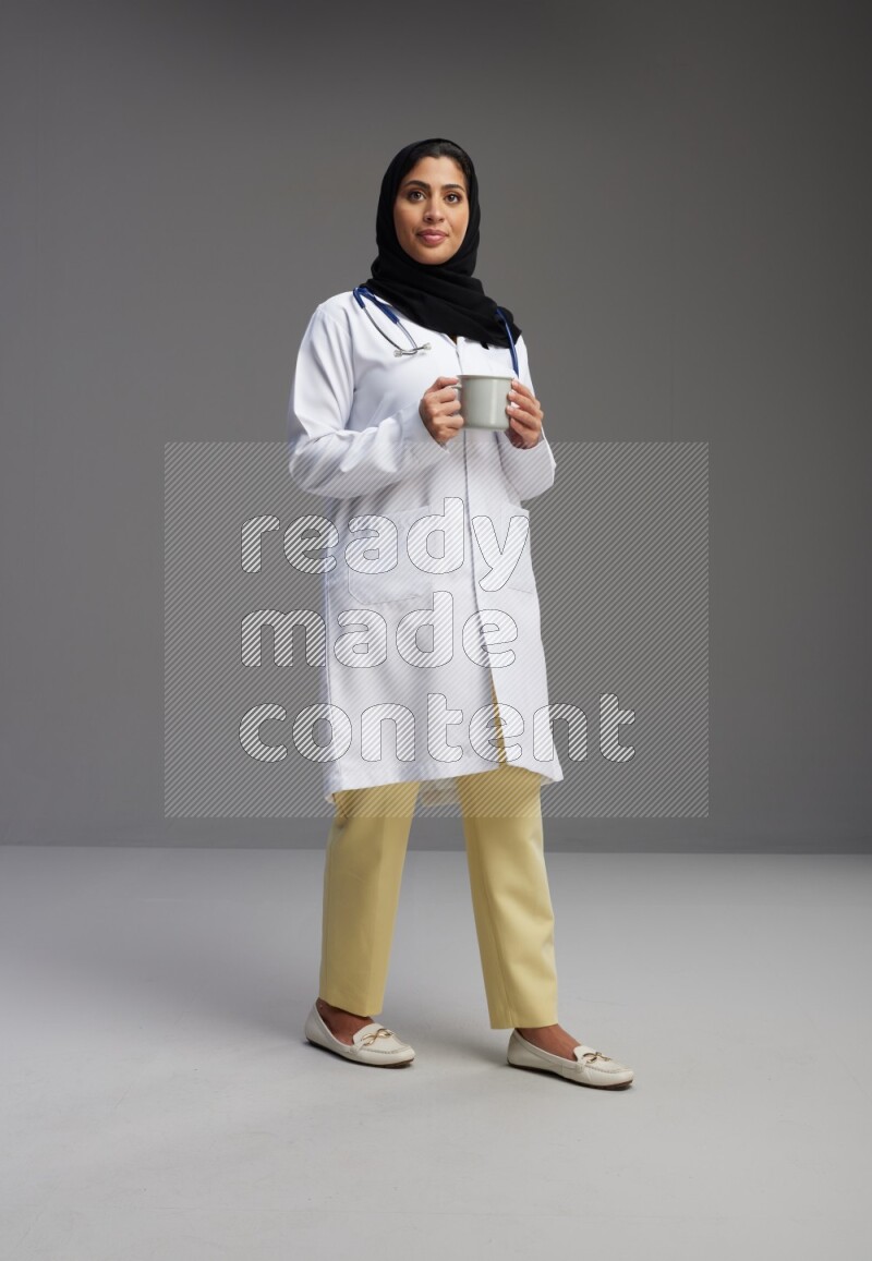 Saudi woman wearing lab coat with stethoscope standing holding mug on Gray background