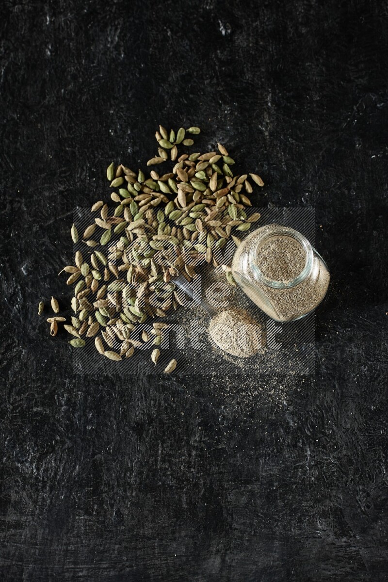 A glass spice jar and a metal spoon full of cardamom powder and cardamom seeds spreaded on textured black flooring