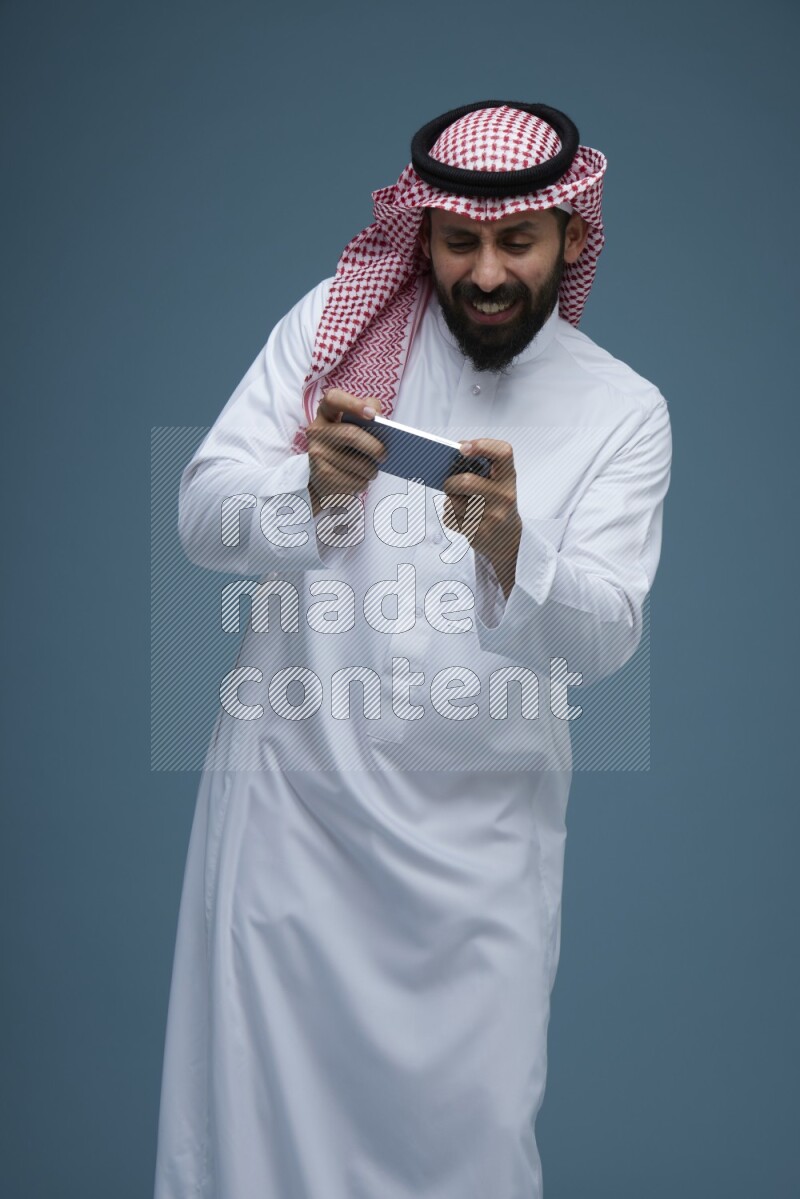 A man Playing a Game on his phone in a blue background wearing Saudi Thob with Shomag