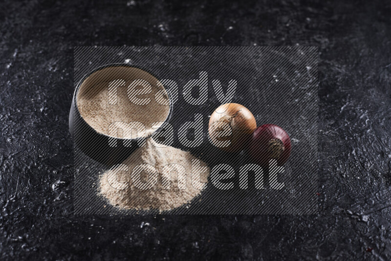 A black pottery bowl full of onion powder with fallen powder from it on black background