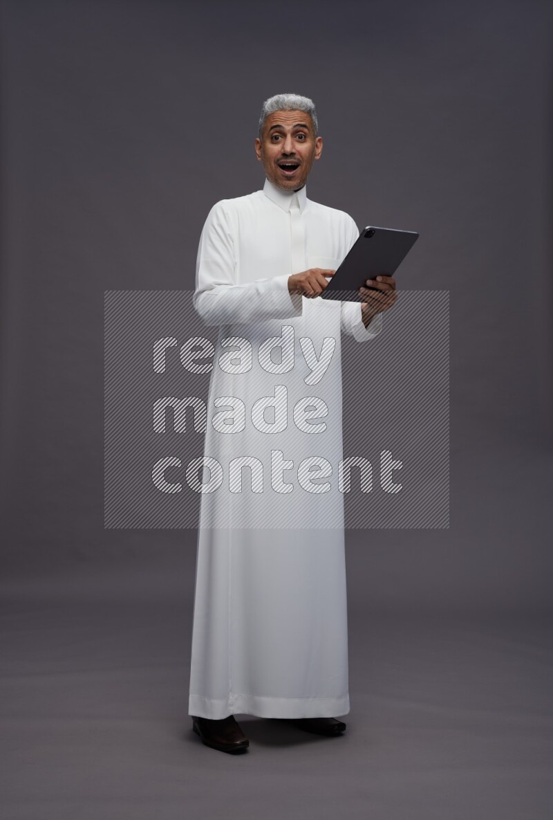 Saudi man wearing thob standing working on tablet on gray background