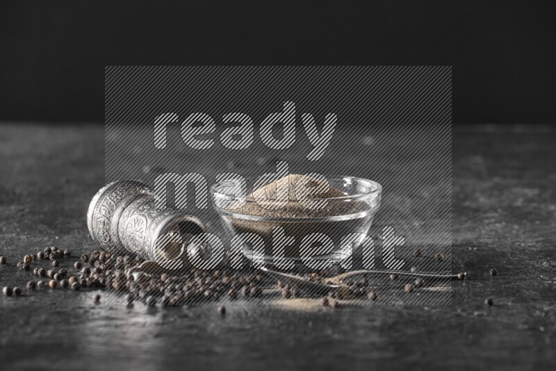 A glass bowl full of black pepper powder with black pepper beads, a turkish metal pepper grinder and a metal spoon on textured black flooring