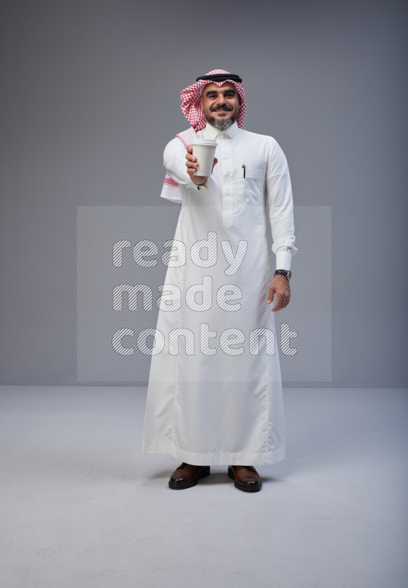 Saudi man Wearing Thob and red Shomag standing holding paper cup on Gray background