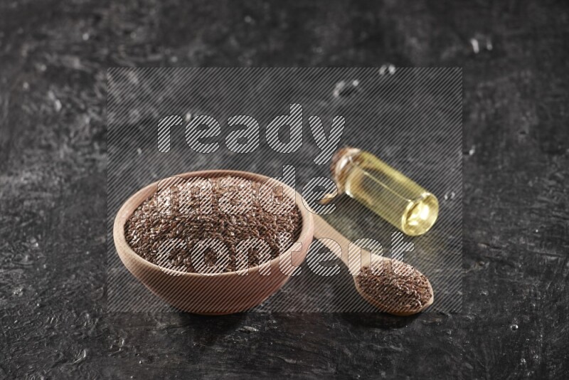 A wooden bowl and spoon full of flaxseeds and a glass bottle of flaxseeds oil on a textured black flooring