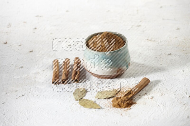 Cinnamon powder in a ceramic bowl with cinnamon sticks and laurel leaves on white background