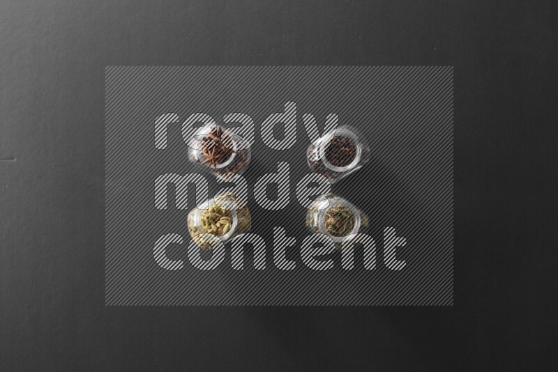 4 glass spice jars full of cloves, cardamom, basil and star anise on black flooring