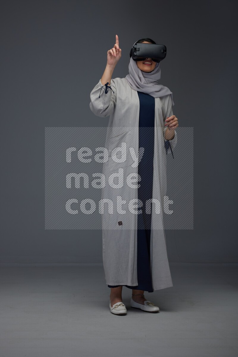 A Saudi woman wearing a light gray Abaya and head scarf standing and wearing VR eye level on a grey background