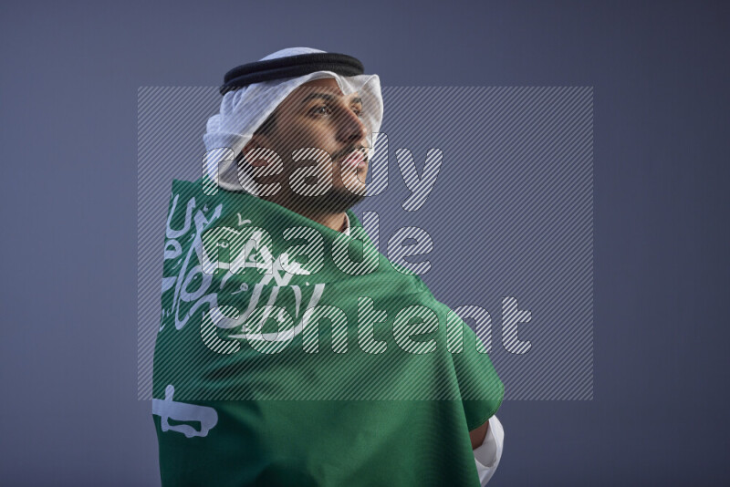 A close-up shot of Saudi man wearing thob and white shomag wrapping big Saudi flag on gray background