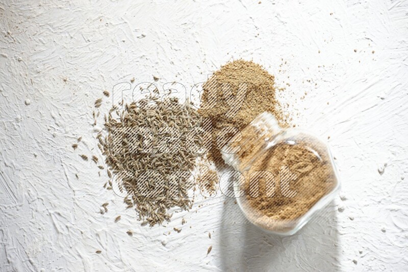 A flipped glass spice jar full of cumin powder and powder spilled out with cumin seeds on textured white flooring