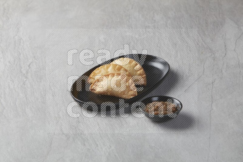 Three fried sambosas in an oval shaped black plate and a sauce in a black round ramekin on a gray background