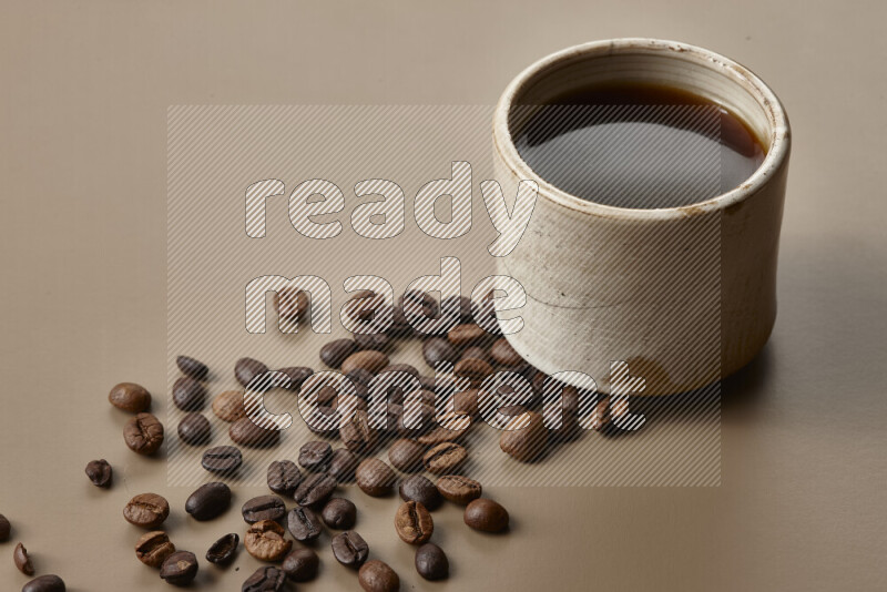 A beige pottery cup of coffee surrounded by roasted coffee beans on beige background