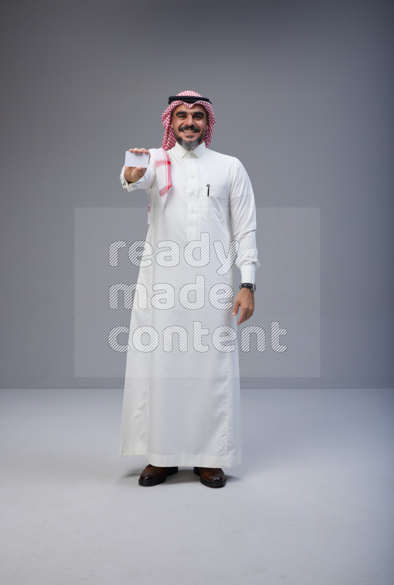 Saudi man Wearing Thob and red Shomag standing holding ATM card on Gray background