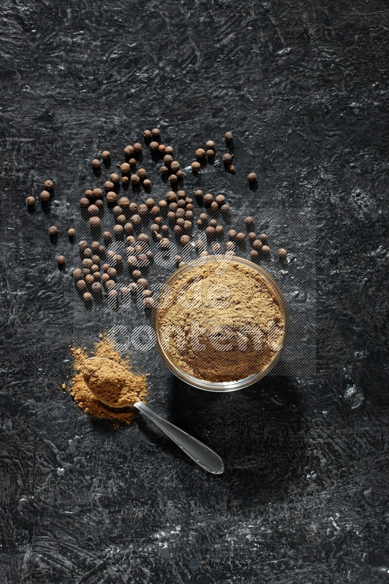 A glass bowl and metal spoon full of allspice powder and whole balls spreaded on a black flooring
