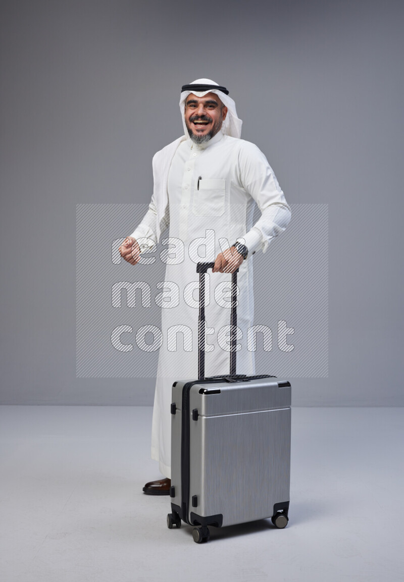 Saudi man wearing Thob and white Shomag standing holding Travel bag on Gray background