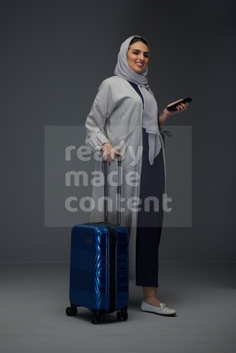 A Saudi woman wearing a light gray Abaya and head scarf standing holding a phone and being shocked  eye level on a grey background