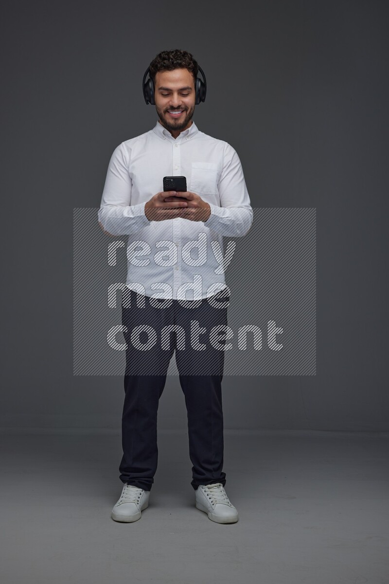 A man wearing smart casual and using his phone and headphone eye level on a gray background