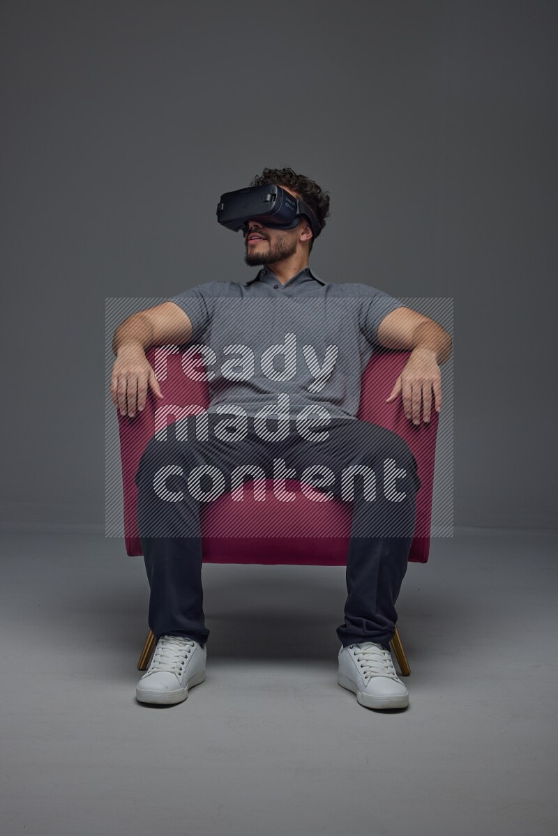A man wearing casual and wearing VR while sitting on a burgundy chair eye level on a gray background