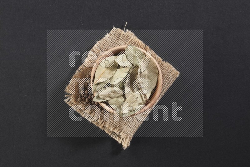 A wooden bowl filled with dried bay leaves on a piece of burlap with bunch of allspice berries on black flooring