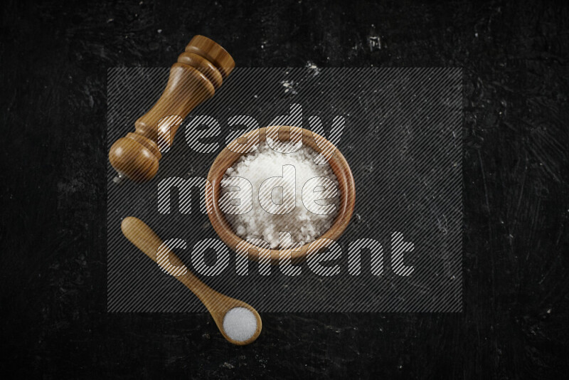 A wooden bowl and spoon filled with white sea salt and wooden grinder beside them on black background