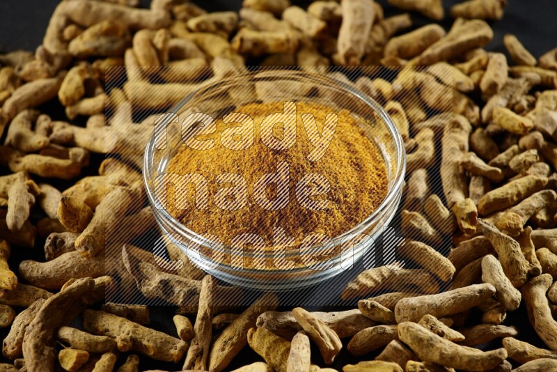 A glass bowl full of turmeric powder and dried whole fingers beside it on black flooring