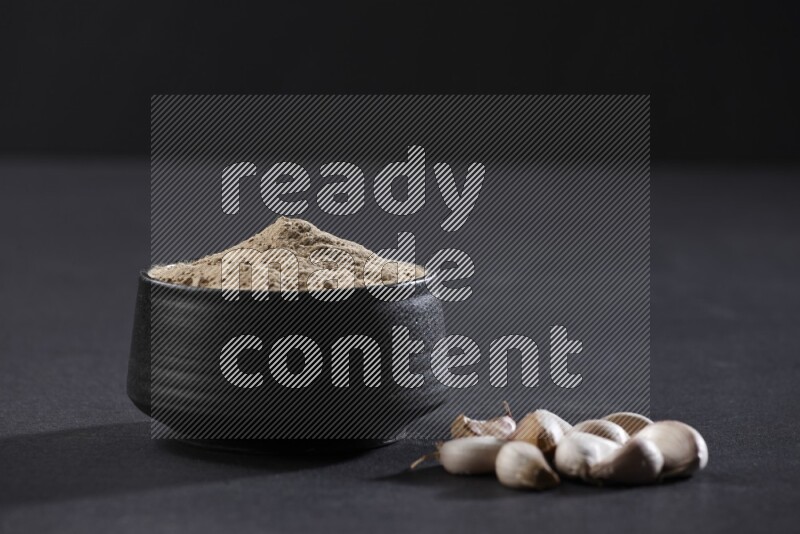 A black pottery bowl full of garlic powder with some cloves beside it on a black flooring