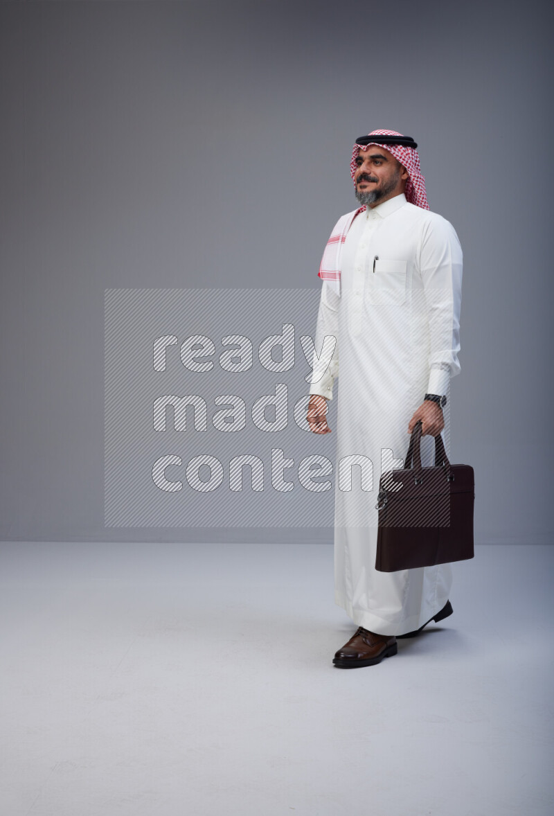 Saudi man Wearing Thob and red Shomag standing holding bag on Gray background