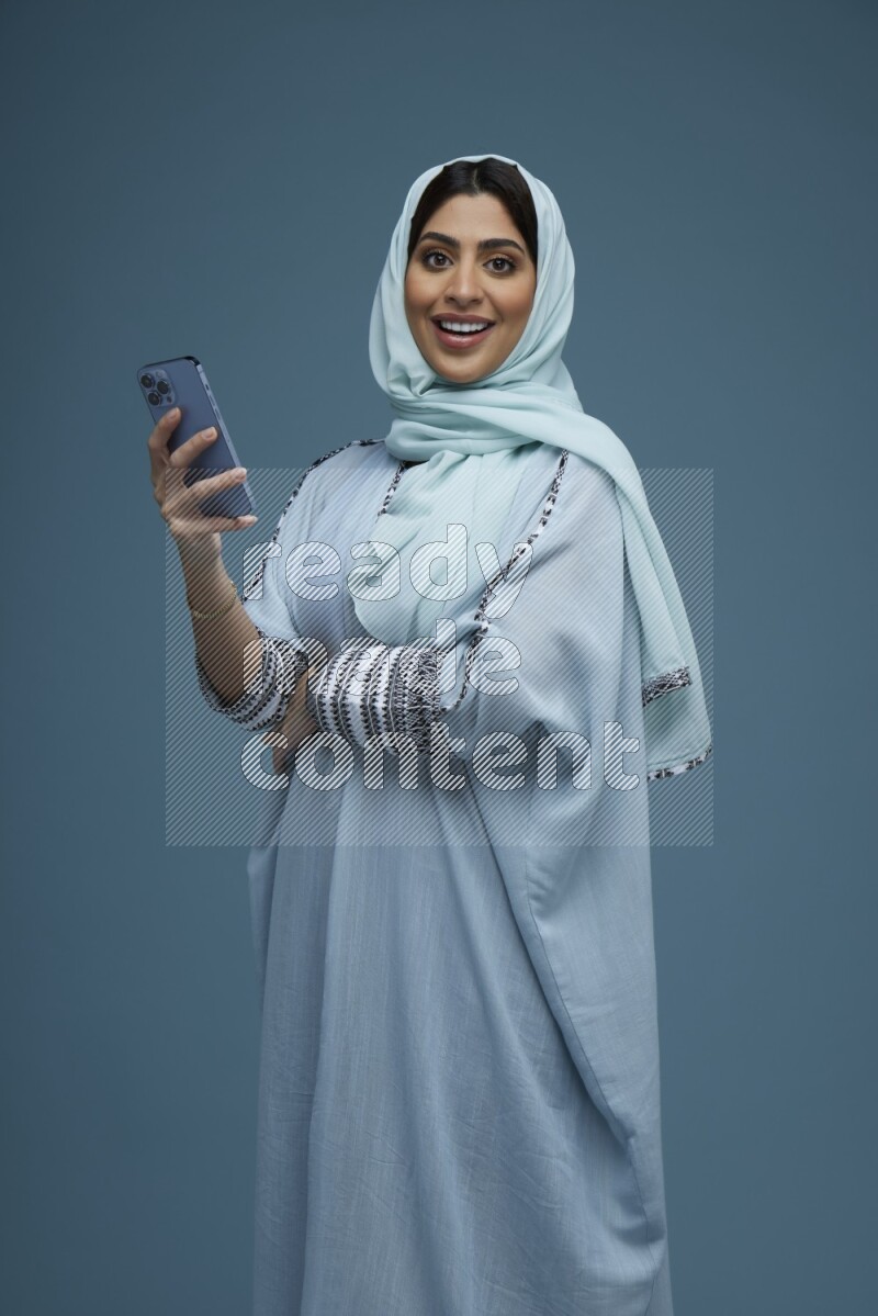 A Saudi woman posing with her phone in a blue background wearing a blue Abaya with hijab