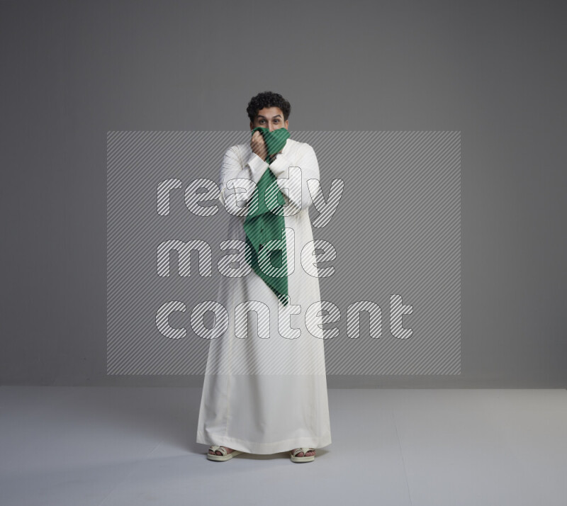 A Saudi man standing wearing thob kissing Saudi flag on gray background