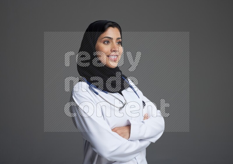 Saudi woman wearing lab coat with stethoscope standing with crossed arms on Gray background