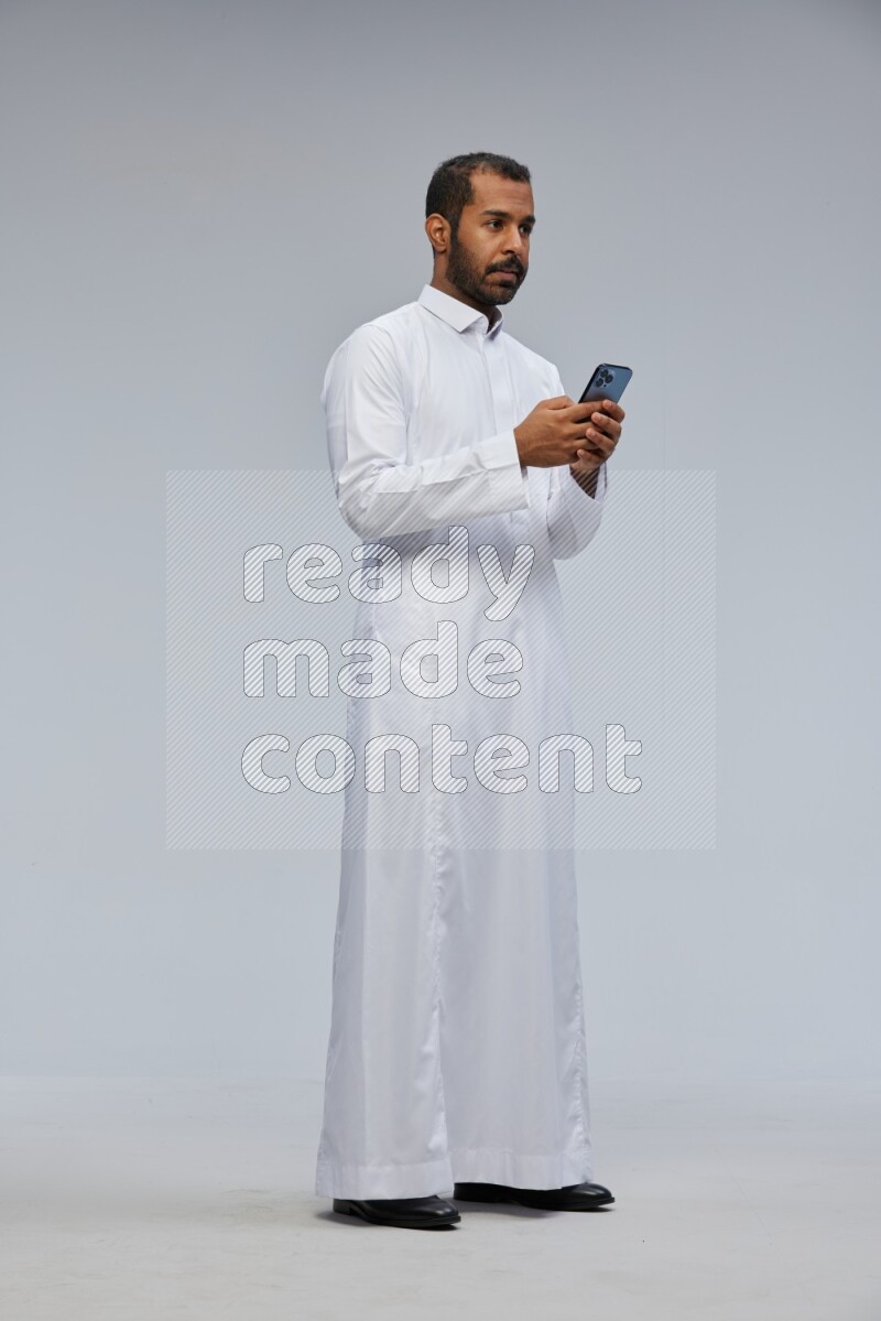 Saudi man wearing Thob standing texting on phone on Gray background