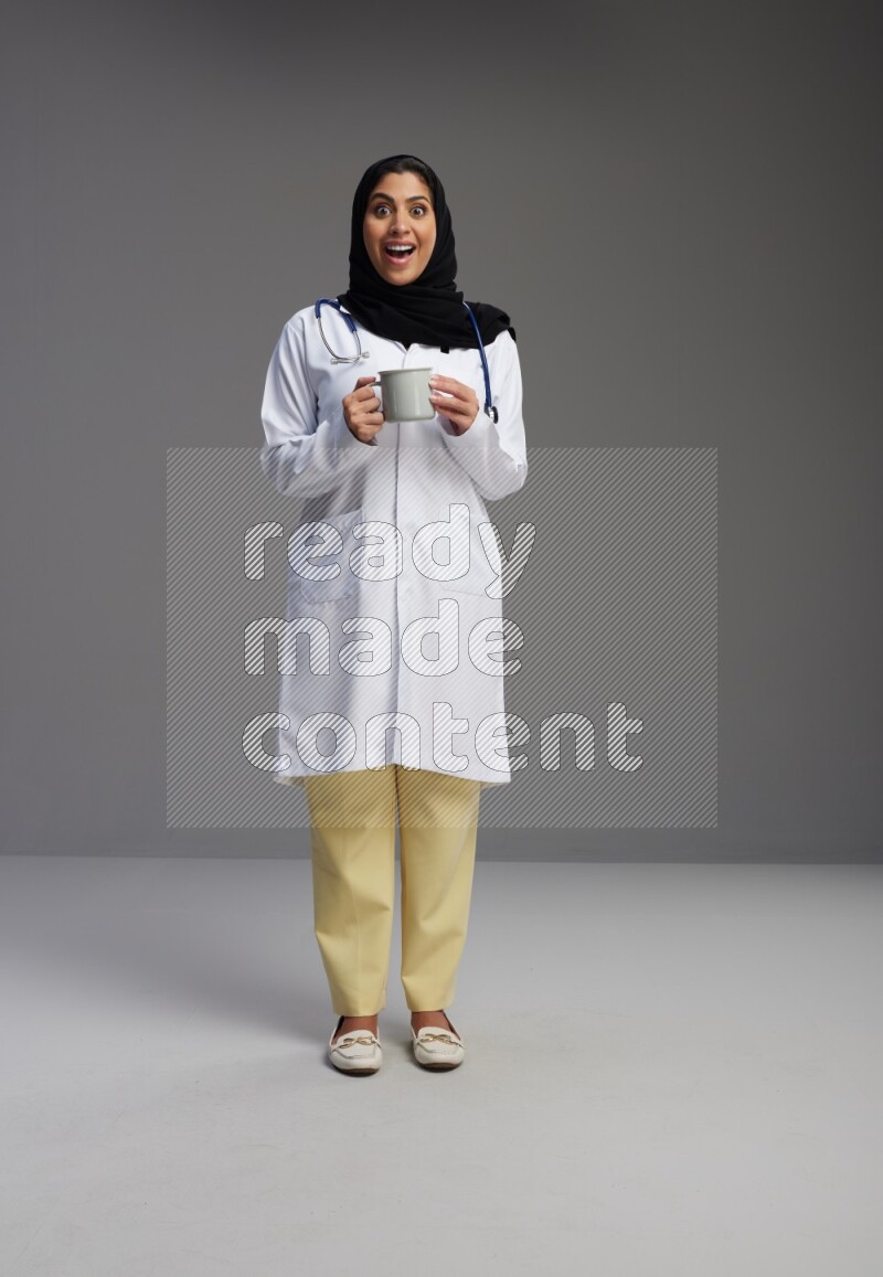 Saudi woman wearing lab coat with stethoscope standing holding mug on Gray background
