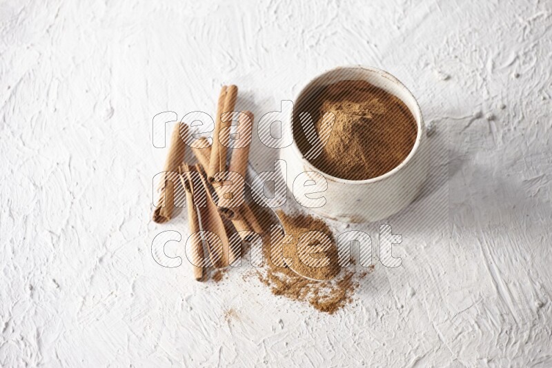 Ceramic beige bowl full of cinnamon powder and a metal spoon with cinnamon sticks next of it on a textured white background