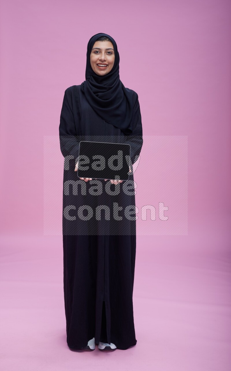 Saudi woman wearing Abaya standing showing tablet to camera on pink background