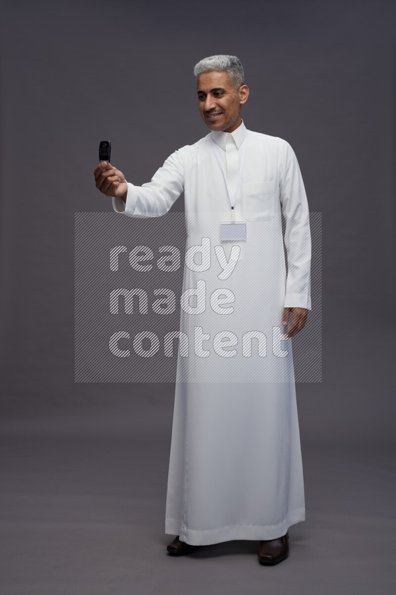 Saudi man wearing thob with neck strap employee badge standing holding key car on gray background