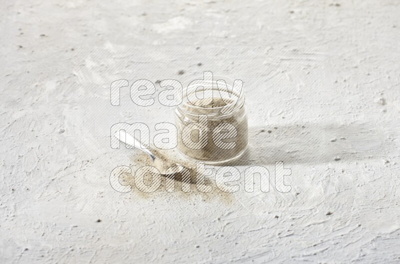 A glass jar and a metal spoon full of white pepper powder on textured white flooring