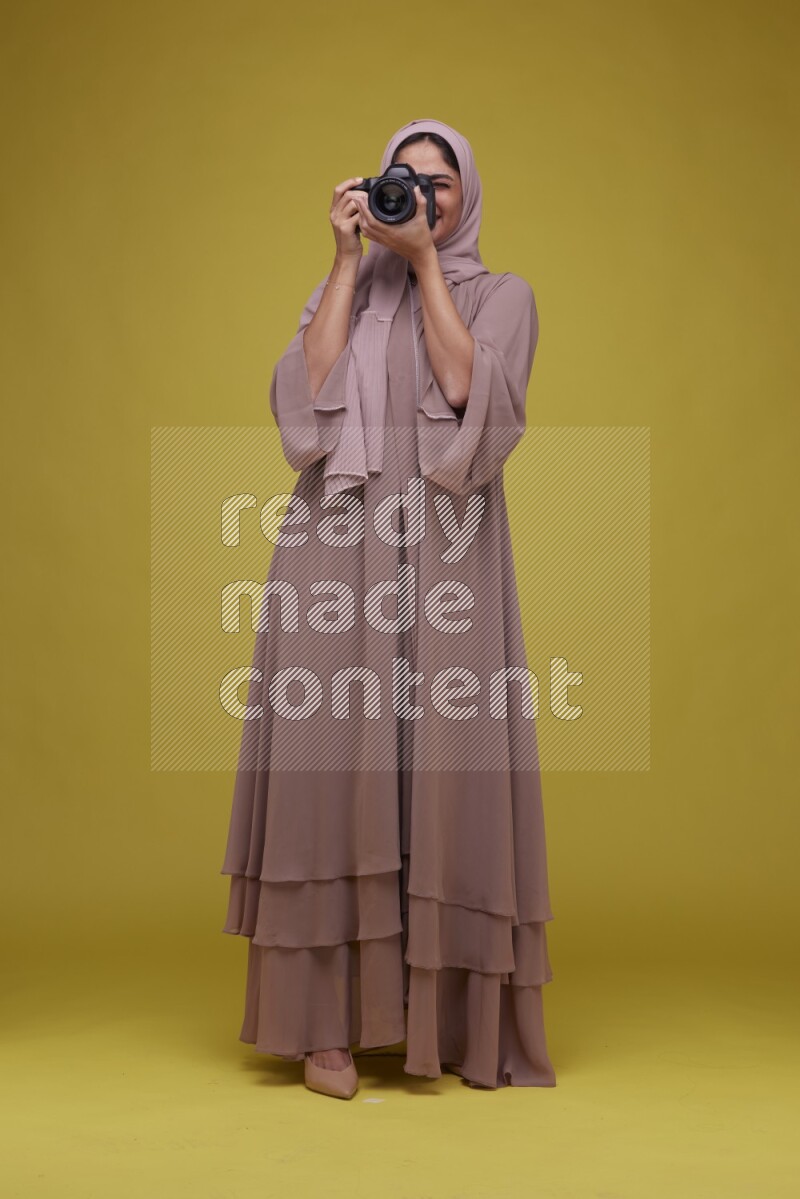 A woman Holding a Camera on a Yellow Background wearing Brown Abaya with Hijab