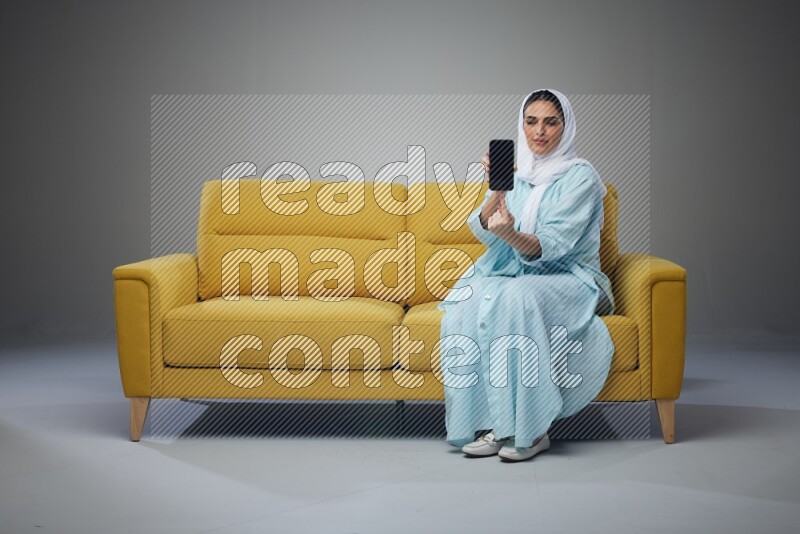 A Saudi woman wearing a light blue Abaya and white head scarf sitting on a yellow sofa and showing her phone's screen vertically and horizontally eye level on a grey background