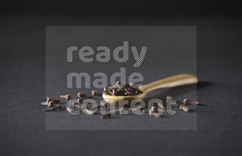 A wooden spoon full of cloves and some spreaded out of it on a black flooring
