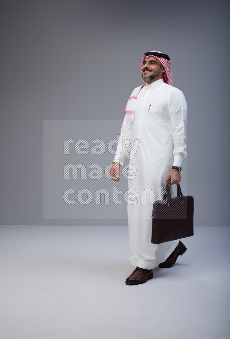 Saudi man Wearing Thob and red Shomag standing holding bag on Gray background