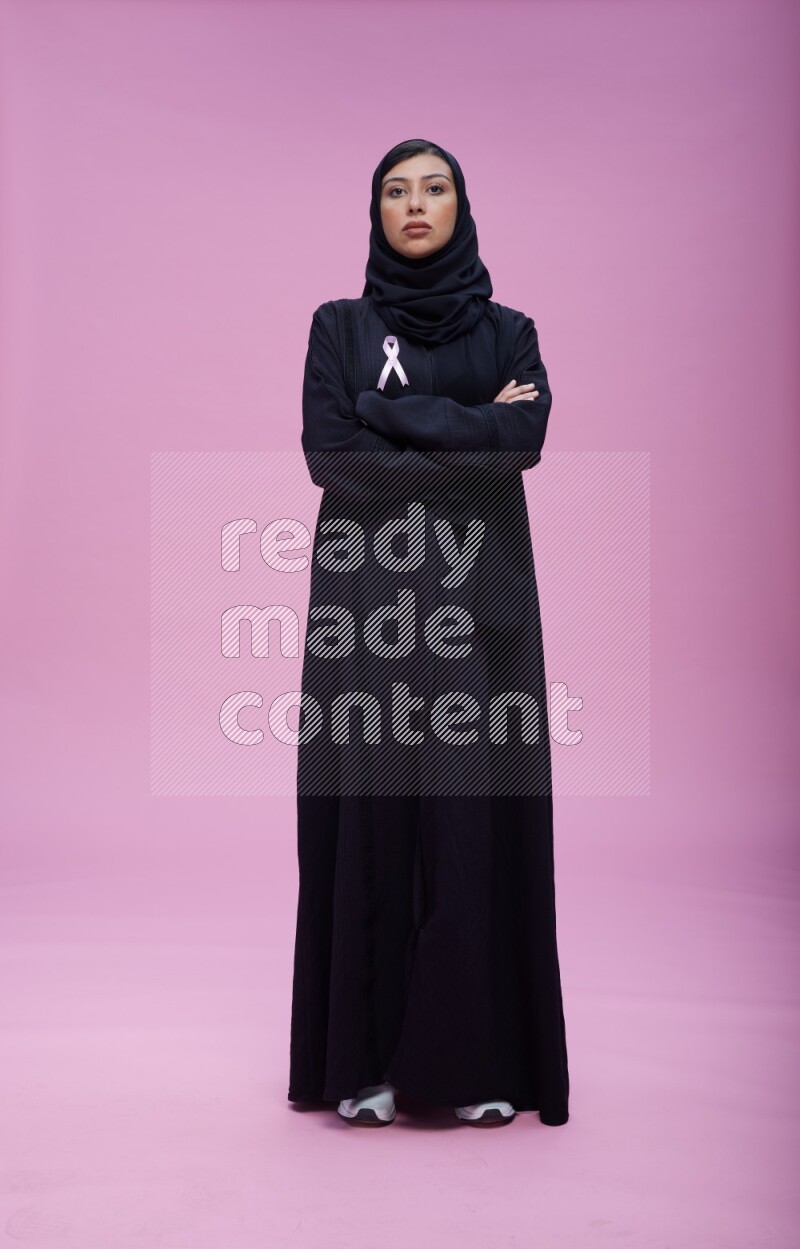 Saudi woman wearing pink ribbon on Abaya standing crossed arms on pink background