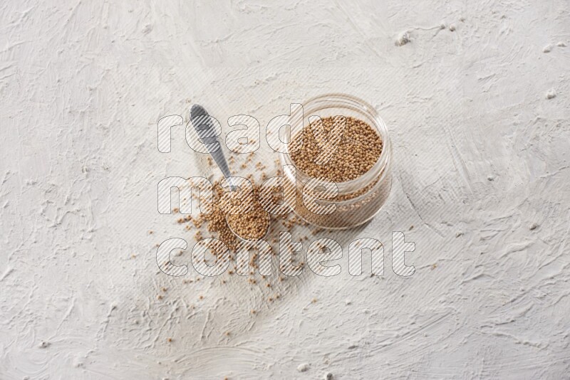 A glass jar and a metal spoon full of mustard seeds on a textured white flooring