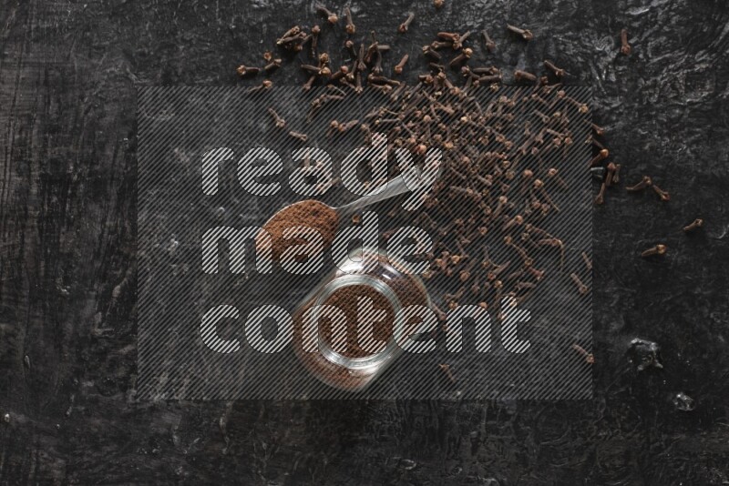 A glass spice jar and a metal spoon full of cloves powder with cloves spread on textured black flooring