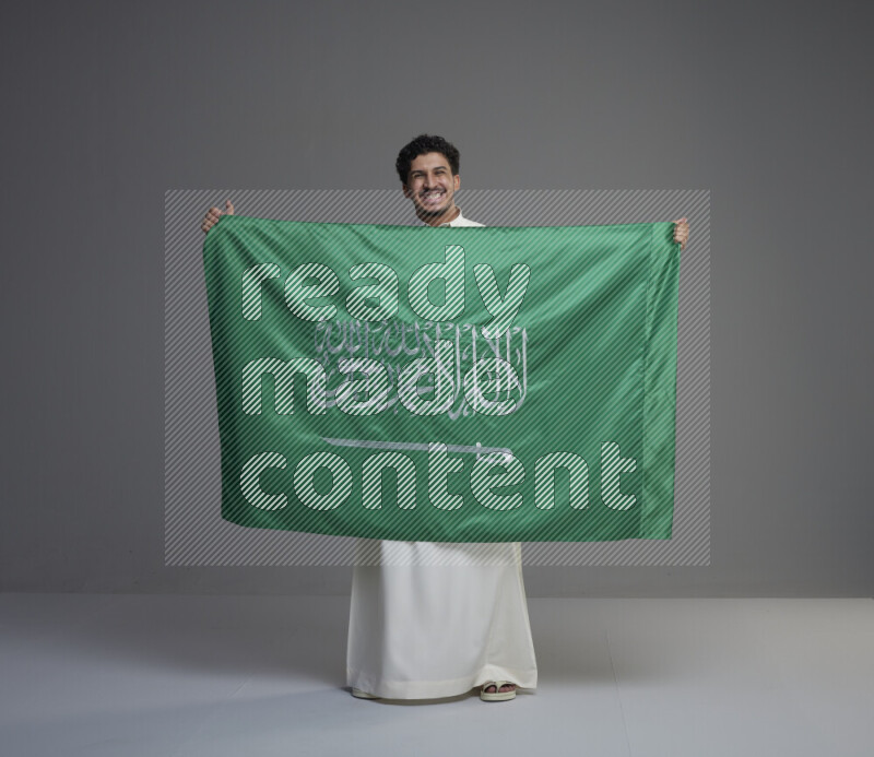A Saudi man standing wearing thob holding big Saudi flag on gray background