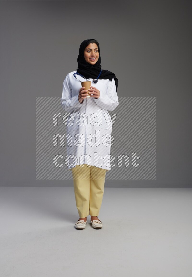 Saudi woman wearing lab coat with stethoscope standing holding paper cup on Gray background