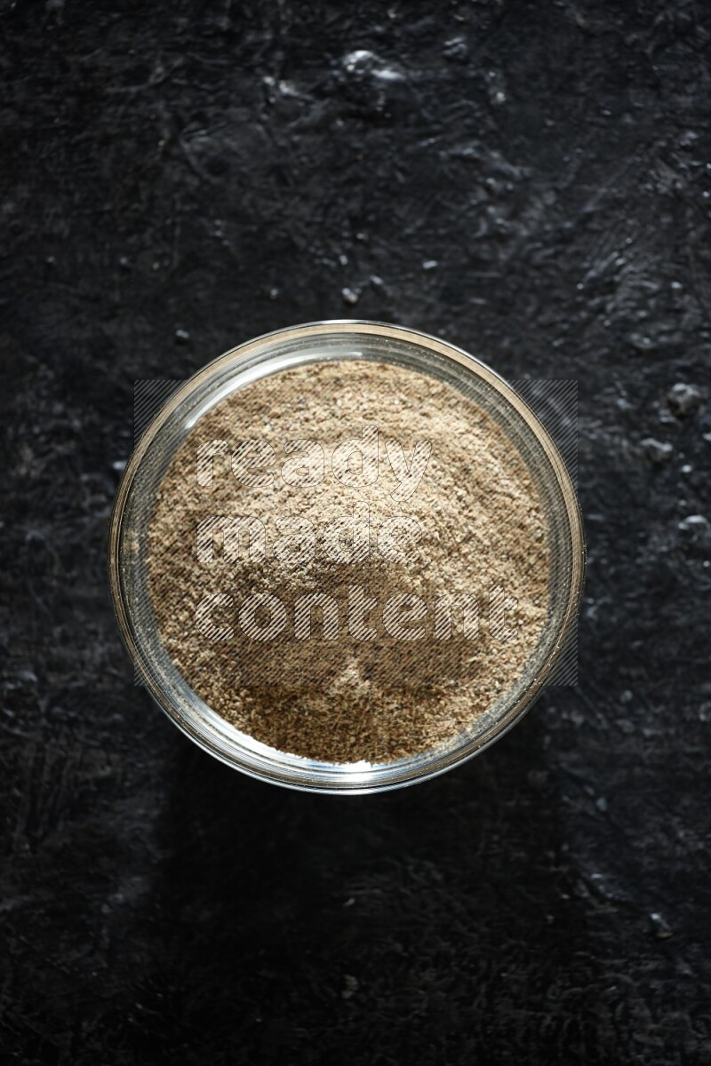 A glass bowl full of cardamom powder on textured black flooring